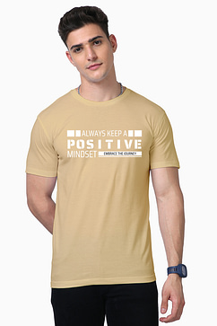 A person in a beige Unisex Supima T-Shirt with Always Keep a Positive Mindset, Embrace the Journey printed on it stands with short dark hair, embodying the laid-back Looga style and posing with one hand in their pocket.