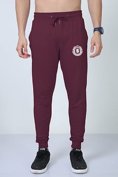 A man wearing Looga's Premium Joggers for Men - Comfort Meets Style, in a sophisticated burgundy color with a white emblem on the upper thigh and an adjustable drawstring, stands against a gray background. Completing his look are black sneakers with white soles and a sleek blue watch.