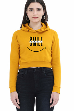 A person wearing a yellow cropped hoodies with SMILE in large letters, a crossed-out word above it, and a smiley face graphic below. They have long hair and are standing against a plain background.