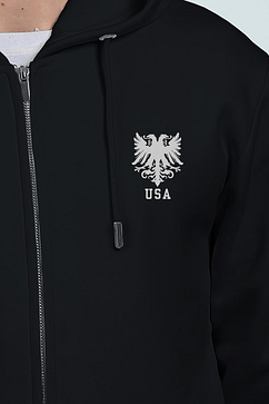 A person is seen from the chest up, wearing a black embroidered hoodie featuring a silver double-headed eagle emblem and USA text.