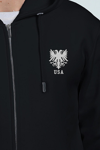 A person is seen from the chest up, wearing a black embroidered hoodie featuring a silver double-headed eagle emblem and USA text.