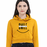 A person wearing a yellow cropped hoodies with SMILE in large letters, a crossed-out word above it, and a smiley face graphic below. They have long hair and are standing against a plain background.